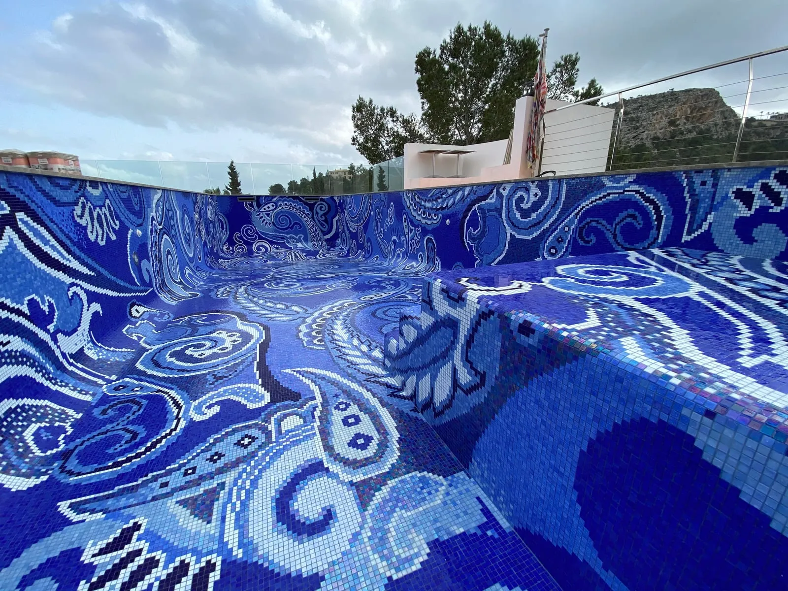 Leeres Außenbecken, von der Beckenwand aus gesehen: das blau-weiße Paisley-Ornament läuft über Boden, Hohlkehle und Wände, dahinter Bäume und ein Bergrücken.