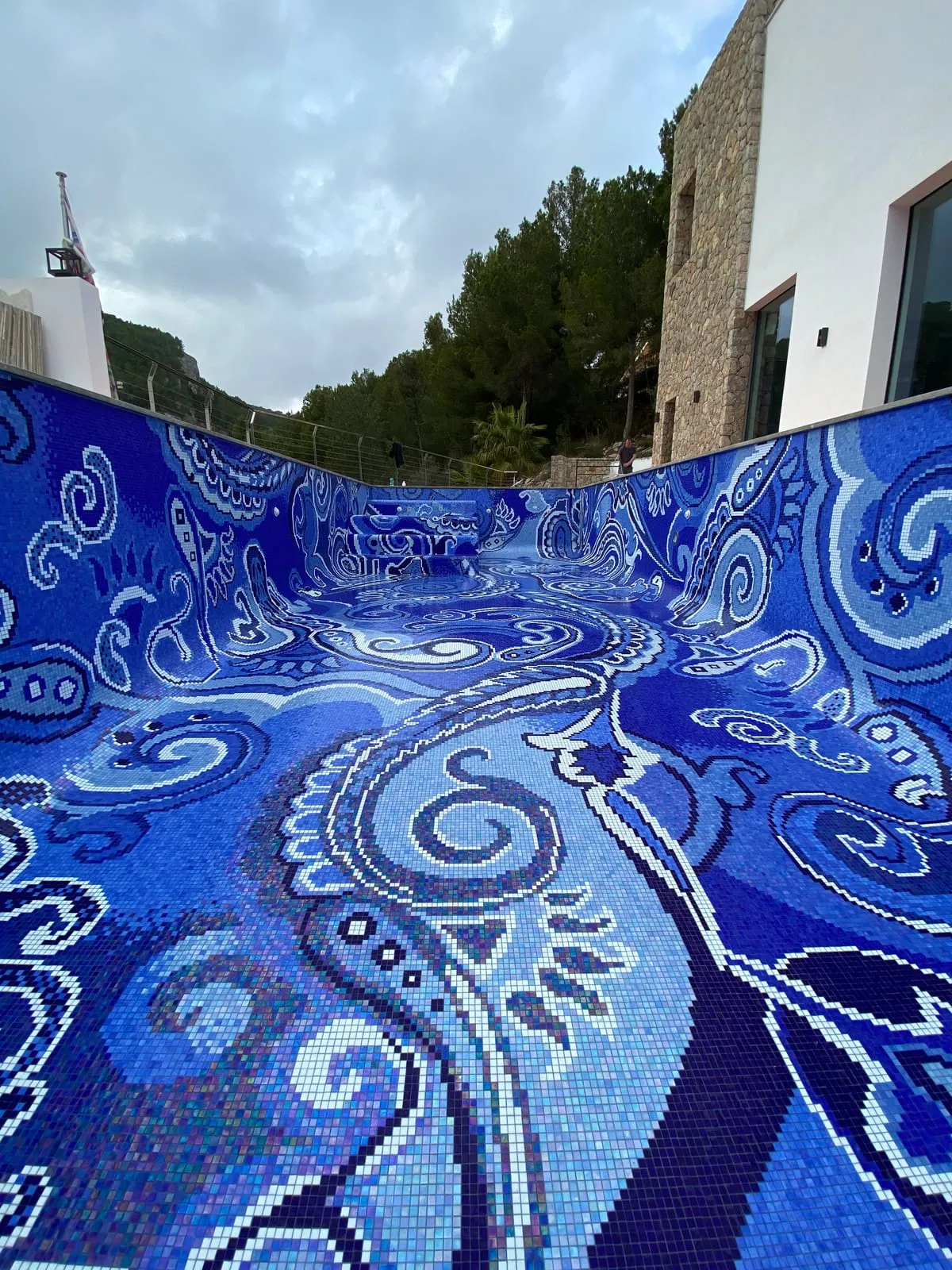 Blick in das leere Becken entlang der langen Wand, das Paisley-Muster zieht über die Rundung vom Boden in die Wand, im Hintergrund das Haus mit Natursteinfassade.