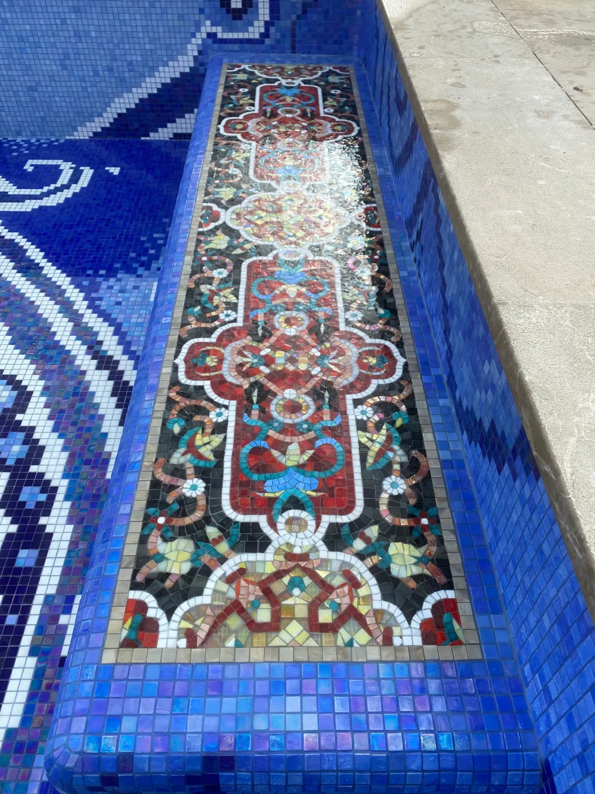 Die Oberseite einer Beckenstufe trägt ein zweites Bildmosaik wie einen Teppichläufer: rotes und cremefarbenes Ornament auf Schwarz, gerahmt von blauem Glasmosaik.