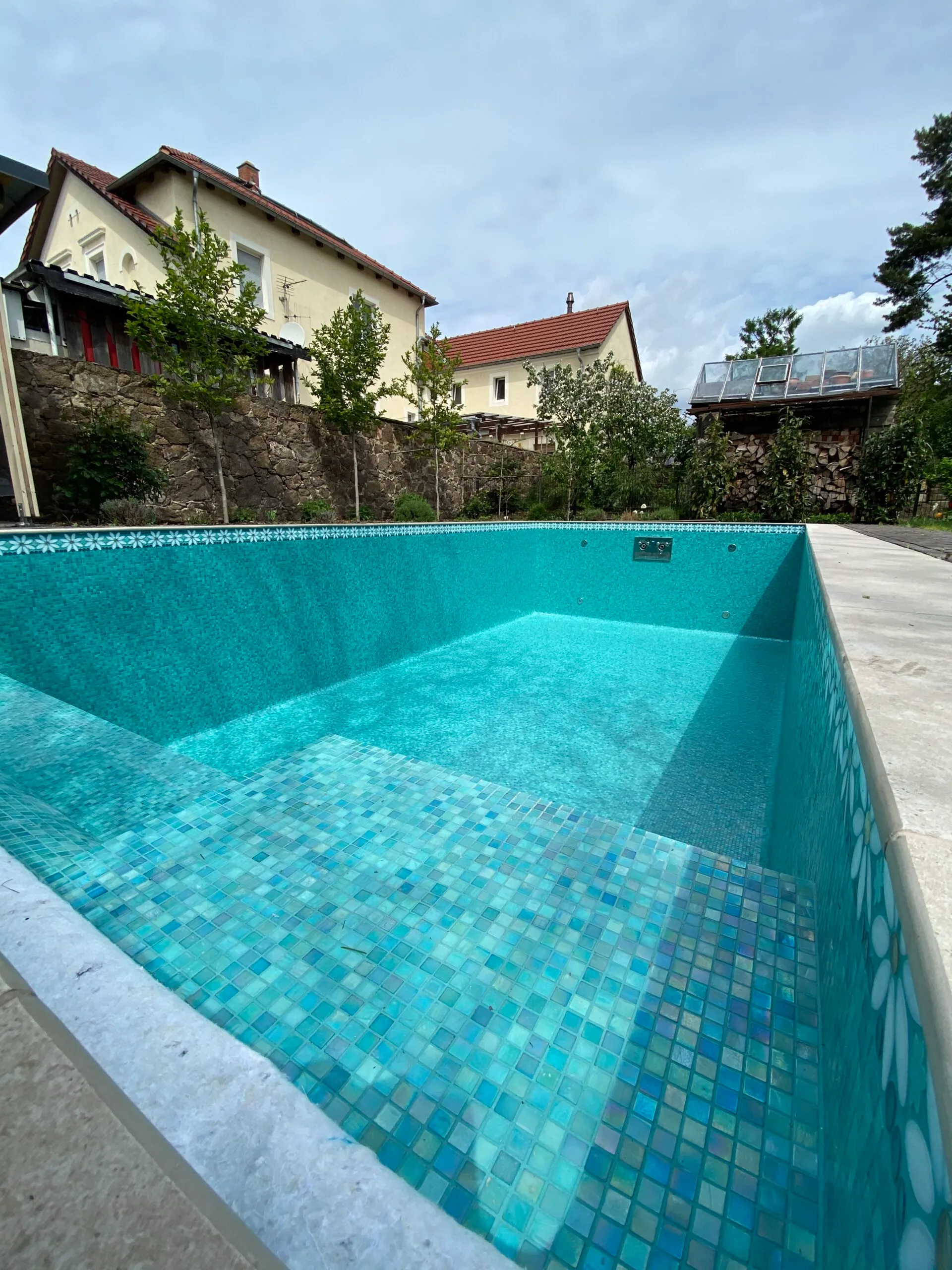 Leeres Gartenbecken mit irisierendem türkisfarbenem Mosaik und einer umlaufenden Bordüre aus weißen Blüten an der Wasserlinie.