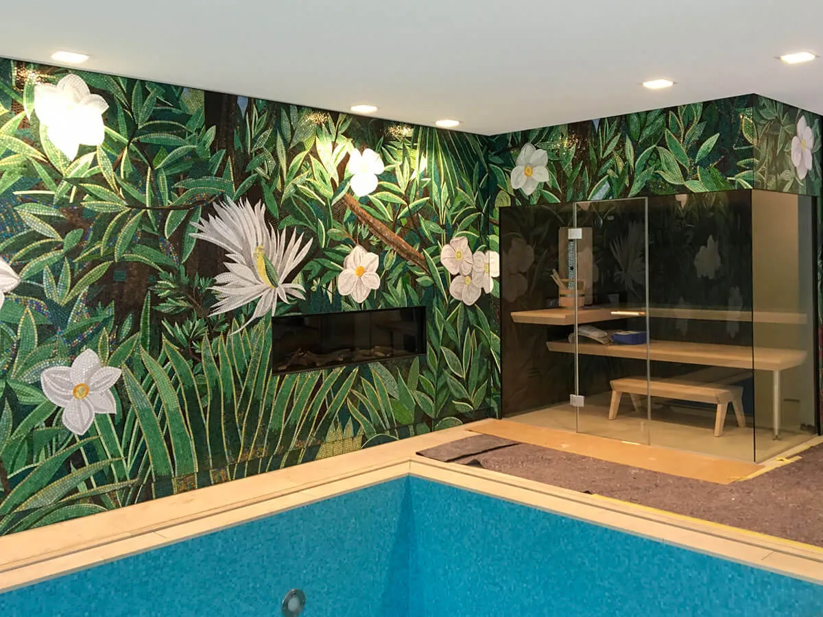 Innenpool, dessen Wandmosaik um die Raumecke weiterläuft und eine Sauna hinter Glas sowie einen wandbündigen Kamin rahmt.