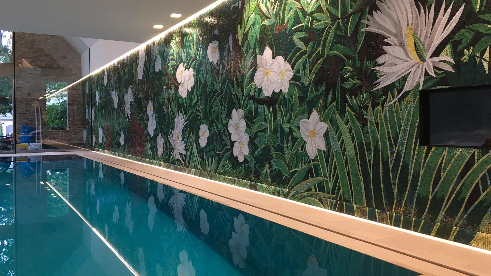 Innenpool mit raumhohem Wandmosaik aus grünen Blättern und weißen Blüten, das sich im türkisfarbenen Wasser spiegelt.